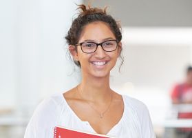 A student in a white blouse smiling while holding a red notebook.
